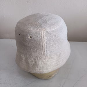 Pode incluir: Um chap&eacute;u de pescador liso em tom branco sujo, feito de tecido felpudo texturizado. O chap&eacute;u apresenta uma copa plana, uma aba inclinada para baixo com detalhes de costura e dois pequenos ilh&oacute;s met&aacute;licos circulares na lateral.