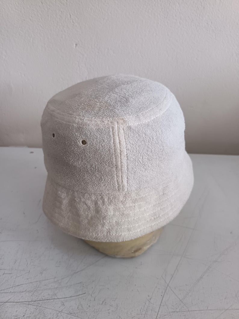 Pode incluir: Um chap&eacute;u de pescador liso em tom branco sujo, feito de tecido felpudo texturizado. O chap&eacute;u apresenta uma copa plana, uma aba inclinada para baixo com detalhes de costura e dois pequenos ilh&oacute;s met&aacute;licos circulares na lateral.