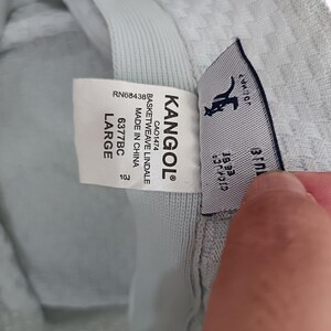 May include: A light gray bucket hat with a textured basketweave pattern. The interior features a white label with the text KANGOL, BASKETWEAVE LINDALE, MADE IN CHINA, 6377BC, LARGE, and RN68438. A second tag displays the Kangol kangaroo logo.