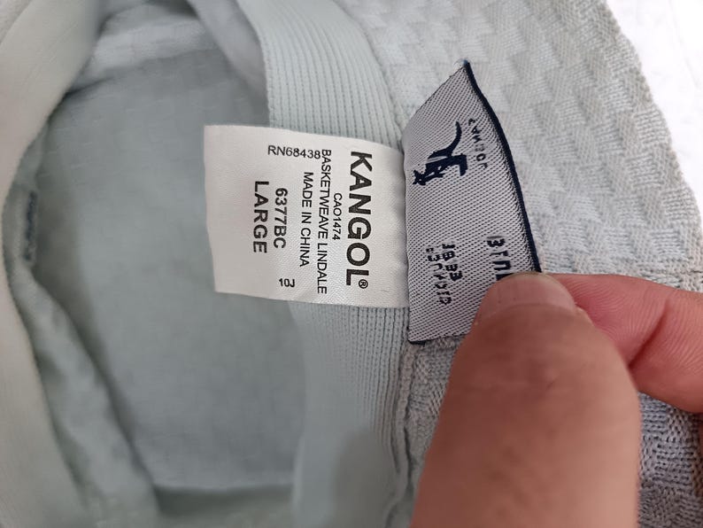 May include: A light gray bucket hat with a textured basketweave pattern. The interior features a white label with the text KANGOL, BASKETWEAVE LINDALE, MADE IN CHINA, 6377BC, LARGE, and RN68438. A second tag displays the Kangol kangaroo logo.