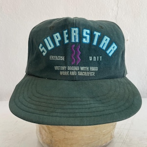 Puede incluir: Un sombrero de pescador verde oscuro con la palabra "SUPERSTAR" bordada en azul claro en la parte delantera. Debajo, se leen las palabras "EXERCISE UNIT" y la frase "VICTORY BEGINS WITH HARD WORK AND SACRIFICE".