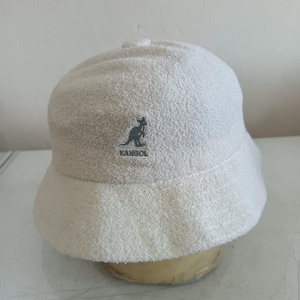 May include: A white bucket hat made of a textured terry cloth material. The front of the hat features an embroidered dark gray logo of a kangaroo silhouette above the text KANGOL. The hat has a rounded crown and a downward sloping brim.