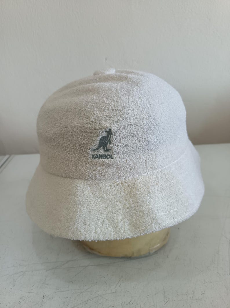 May include: A white bucket hat made of a textured terry cloth material. The front of the hat features an embroidered dark gray logo of a kangaroo silhouette above the text KANGOL. The hat has a rounded crown and a downward sloping brim.