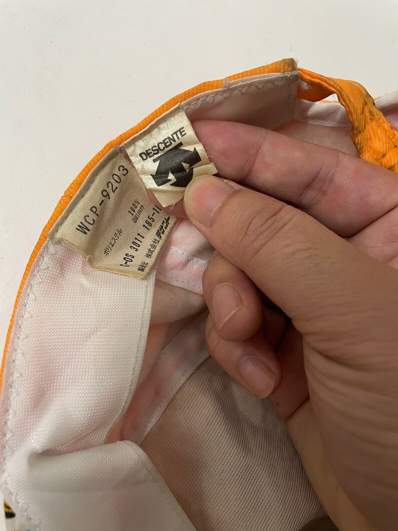 May include: A white and orange baseball cap with a white label that says "WCP-9203 DESCENTE" and has the text "ポリエステル 100% (021002) -OS 3011 185-11 製株式会社デ" in Japanese.