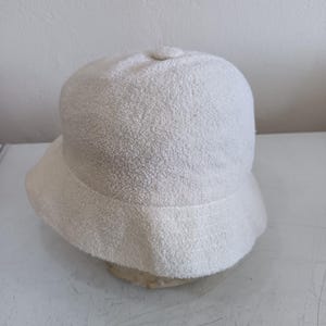 May include: A solid off-white bucket hat featuring a textured, terry cloth-like fabric surface. The hat has a rounded crown with a small central button detail and a downward-sloping brim that encircles the entire circumference of the headpiece.