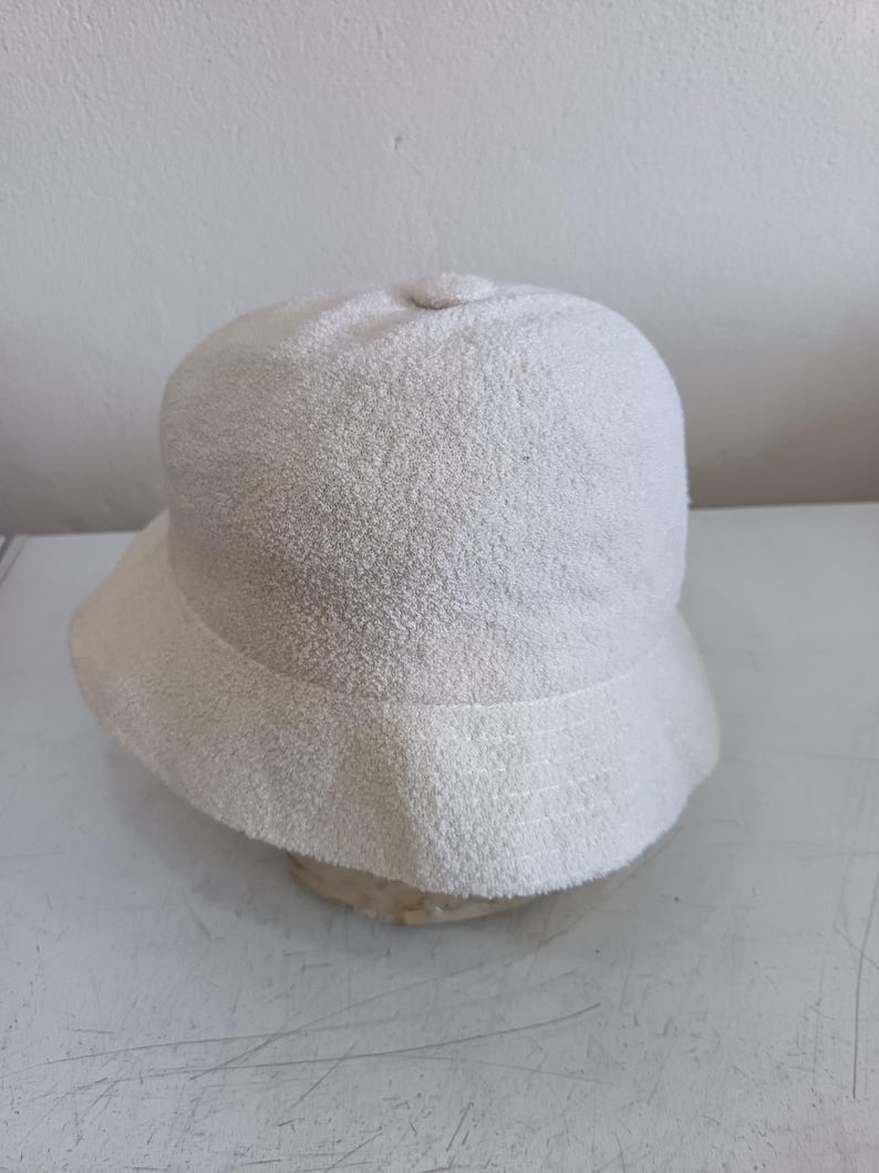 May include: A solid off-white bucket hat featuring a textured, terry cloth-like fabric surface. The hat has a rounded crown with a small central button detail and a downward-sloping brim that encircles the entire circumference of the headpiece.