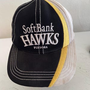 May include: A black, tan, and yellow baseball cap with the text "SoftBank HAWKS" embroidered in white. The cap also features the word "FUKUOKA" and white stitching.