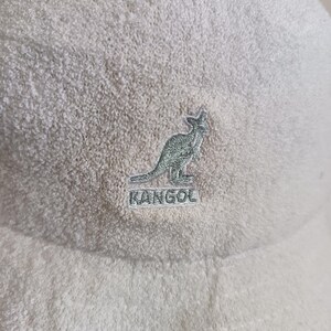 May include: A cream-colored bucket hat made of a textured terry cloth fabric. The front of the hat features an embroidered logo of a kangaroo silhouette positioned above the word KANGOL, both stitched in a light green thread on the fabric.