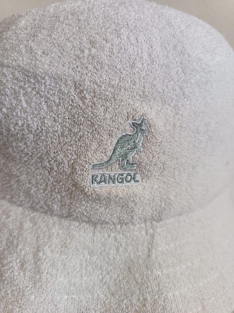 May include: A cream-colored bucket hat made of a textured terry cloth fabric. The front of the hat features an embroidered logo of a kangaroo silhouette positioned above the word KANGOL, both stitched in a light green thread on the fabric.