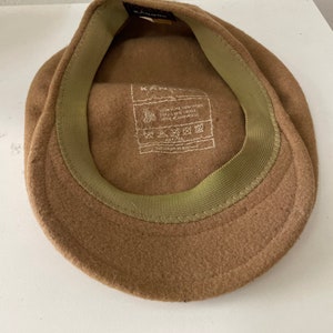 以下が含まれることがあります： 内側に緑色の布製のバンドが付いた、茶色のフェルト製のニュースボーイキャップ。キャップの内側のラベルには「Kangol 100% Pure New Wool Made in England」と記載されています。