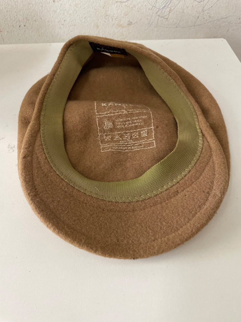 以下が含まれることがあります： 内側に緑色の布製のバンドが付いた、茶色のフェルト製のニュースボーイキャップ。キャップの内側のラベルには「Kangol 100% Pure New Wool Made in England」と記載されています。