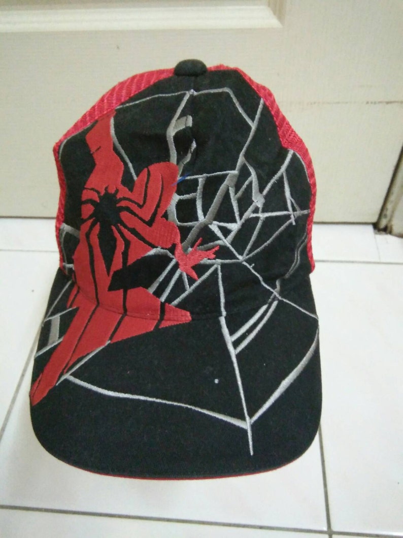 May include: Black and red baseball cap with a spider web design and a red Spiderman figure. The cap has a mesh back.