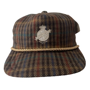 Könnte beinhalten: Eine braune und schwarze karierte Baseballkappe mit goldener Kordel. Die Kappe hat ein weiß gesticktes Logo mit der Aufschrift "Borsalino" und einer Krone über dem Wort.