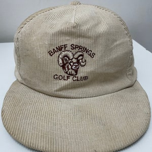May include: A beige corduroy baseball cap with a brown embroidered logo that says "Banff Springs Golf Club" and features a mountain goat.