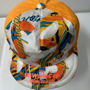May include: A white and orange baseball cap with a colorful abstract design. The cap has a black and white '92 FIS World Cup Morioka Shizuikuishi' logo on the front.