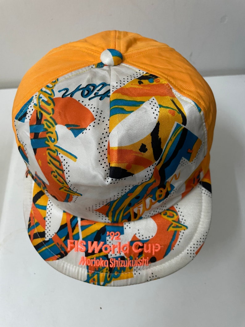 May include: A white and orange baseball cap with a colorful abstract design. The cap has a black and white '92 FIS World Cup Morioka Shizuikuishi' logo on the front.
