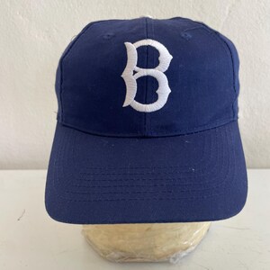 May include: Navy blue baseball cap with a white embroidered "B" logo on the front. The cap has a curved brim and a structured crown. A classic style, suitable for sports or casual wear.