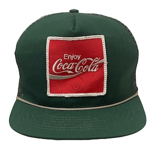 May include: A green trucker hat with a red and white Coca-Cola logo patch. The patch has the words "Enjoy Coca-Cola" on it.