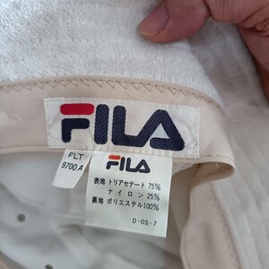 Pode incluir: Um chap&eacute;u bucket de tecido felpudo branco com uma etiqueta da marca FILA no interior. A etiqueta exibe o log&oacute;tipo FILA, o n&uacute;mero de modelo FLT 9700 A e detalhes da composi&ccedil;&atilde;o do tecido em japon&ecirc;s: 75% triacetato, 25% nylon e forro 100% poli&eacute;ster.