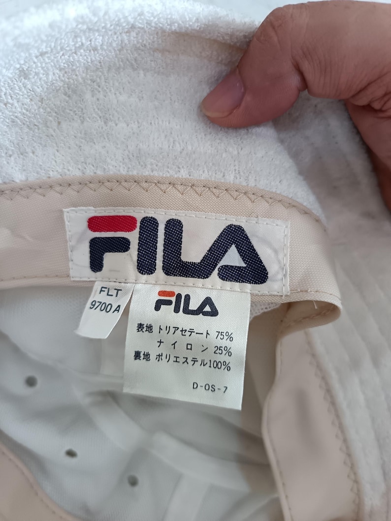 Pode incluir: Um chap&eacute;u bucket de tecido felpudo branco com uma etiqueta da marca FILA no interior. A etiqueta exibe o log&oacute;tipo FILA, o n&uacute;mero de modelo FLT 9700 A e detalhes da composi&ccedil;&atilde;o do tecido em japon&ecirc;s: 75% triacetato, 25% nylon e forro 100% poli&eacute;ster.