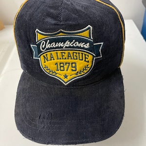 May include: A black corduroy baseball cap with a yellow brim and a yellow and blue embroidered patch that says "Champions NA League 1879".