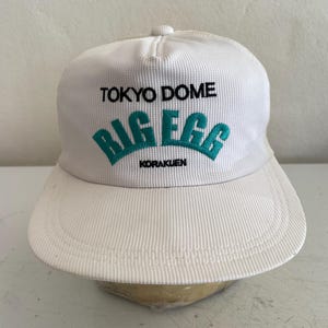 May include: White baseball cap with a curved brim. The cap features the text "TOKYO DOME" above "BIG EGG" in teal, with "KORAKUEN" below. The cap has a textured, ribbed fabric.