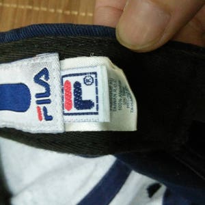 May include: A blue and white baseball cap with a Fila logo on the front. The logo is white with a red outline and a blue "F" inside a white square. The cap has a white label on the side with the text "Fila" and "Made in China" in black.