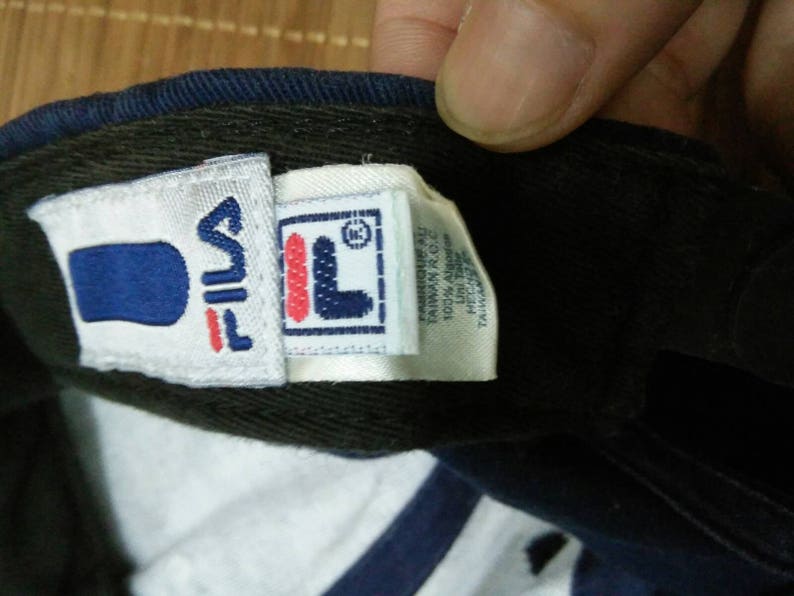 May include: A blue and white baseball cap with a Fila logo on the front. The logo is white with a red outline and a blue "F" inside a white square. The cap has a white label on the side with the text "Fila" and "Made in China" in black.