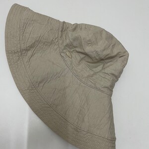 Può includere: Un cappello da sole beige con un'ampia tesa e un piccolo bottone ovale sulla parte superiore.