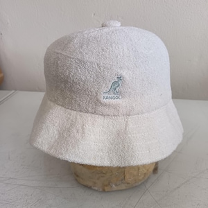 May include: A cream-colored bucket hat made of a textured terry cloth fabric. The front of the hat features an embroidered Kangol logo in a light blue thread, depicting a kangaroo silhouette above the brand name text.