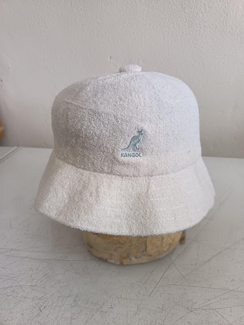 May include: A cream-colored bucket hat made of a textured terry cloth fabric. The front of the hat features an embroidered Kangol logo in a light blue thread, depicting a kangaroo silhouette above the brand name text.