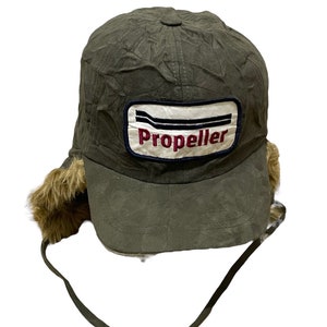 May include: A green faux fur trapper hat with a black and white patch that says "Propeller" in red lettering. The hat has ear flaps that tie at the chin.