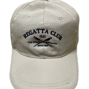 May include: A beige baseball cap with a navy blue embroidered logo that says "REGATTA CLUB" with crossed oars and a small Union Jack flag. The text below the logo says "The Greatest Championship Since 1899."