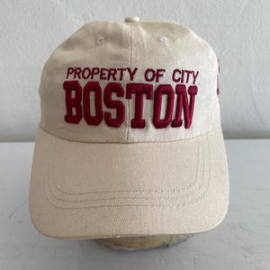 May include: A beige baseball cap with the text "PROPERTY OF CITY BOSTON" embroidered in red. The cap has a curved brim and a button on top. The hat is made of a soft fabric.