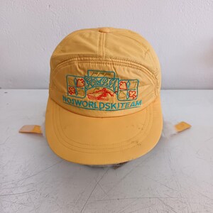 May include: A yellow baseball cap with ear flaps featuring an embroidered graphic of a skier and the text "GOAL" and "NO1WORLDSKITEAM" in teal, with red and orange card suit symbols including hearts, diamonds, clubs, and spades on the front panel.