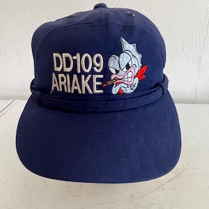 May include: Navy blue baseball cap with the text "DD109 ARIAKE" embroidered in white. The cap features an embroidered cartoon fish with a red scarf and a cigarette. The cap has a curved brim and a button on top.