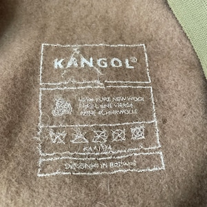 以下が含まれることがあります： Kangolブランドの茶色の帽子。白いラベルには「Kangol」と書いてあり、お手入れ方法が記載されています。ラベルには「Designed in Britain」とも書かれています。