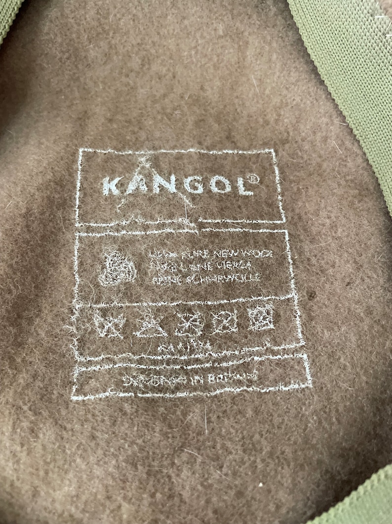 以下が含まれることがあります： Kangolブランドの茶色の帽子。白いラベルには「Kangol」と書いてあり、お手入れ方法が記載されています。ラベルには「Designed in Britain」とも書かれています。