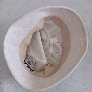 Pode incluir: Um chap&eacute;u bucket de tecido felpudo branco com forro interior bege e coroa de malha branca. O interior apresenta uma etiqueta da marca Fila, uma etiqueta de tamanho M e uma etiqueta de cuidados. O chap&eacute;u tem uma aba larga e estruturada.