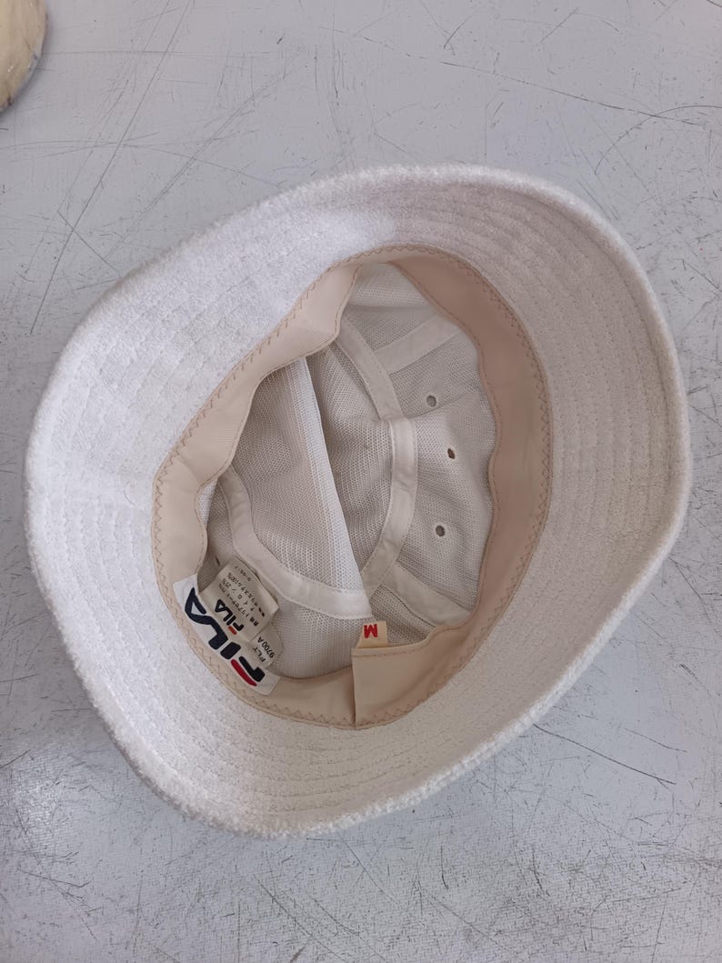 Pode incluir: Um chap&eacute;u bucket de tecido felpudo branco com forro interior bege e coroa de malha branca. O interior apresenta uma etiqueta da marca Fila, uma etiqueta de tamanho M e uma etiqueta de cuidados. O chap&eacute;u tem uma aba larga e estruturada.