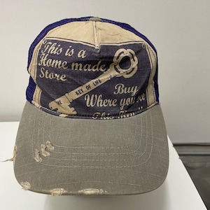 Może przedstawiać: Czapka truckerowa z siatki w kolorze niebieskim i beżowym z przetartym szarym daszkiem. Czapka zawiera reklamę w stylu vintage dla "Home Made Store" z kluczem i tekstem "Buy Where You See This Key".