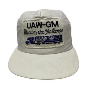 Può includere: Cappellino da camionista bianco con una toppa ricamata in nero e blu che dice "UAW-GM Meeting the Challenge" e presenta un camioncino blu con un rimorchio bianco.