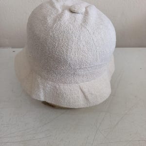 May include: A solid off-white bucket hat with a textured, fuzzy fabric finish. The hat features a rounded crown with a small fabric-covered button at the top center and a downward-sloping brim that encircles the entire base of the hat.