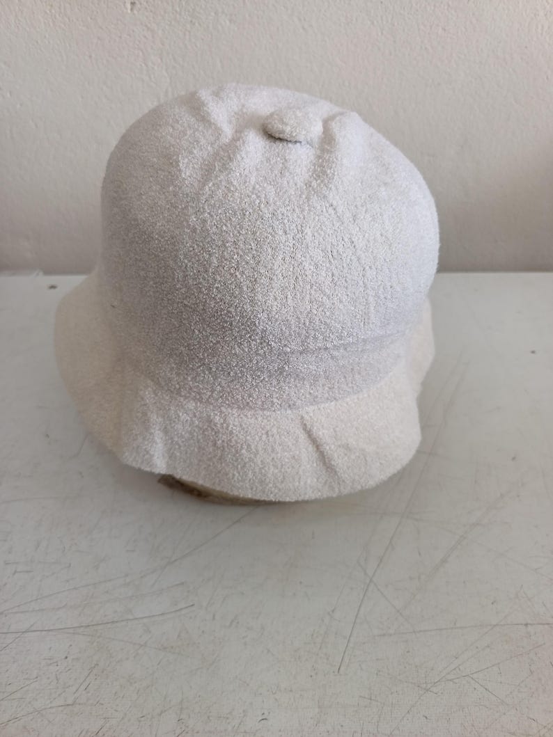 May include: A solid off-white bucket hat with a textured, fuzzy fabric finish. The hat features a rounded crown with a small fabric-covered button at the top center and a downward-sloping brim that encircles the entire base of the hat.