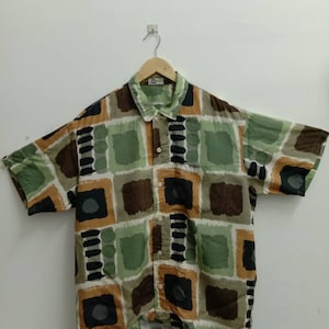 Könnte beinhalten: Ein kurzärmeliges Hemd mit Knöpfen und einem geometrischen Muster in Grün-, Braun- und Schwarztönen. Das Hemd hat einen Kragen und eine Knopfleiste vorne.