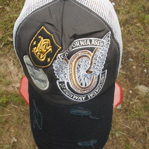 May include: A black and white mesh trucker hat with a gray crown. The hat has a gold embroidered patch with the text "CALIFORNIA ASSN OF HIGHWAY PATROL" and a stylized motorcycle wheel.