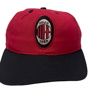 Puede incluir: Una gorra de béisbol roja y negra con el logotipo del AC Milan. El logotipo es un escudo rojo y blanco con el texto "Milan 1899" en blanco.