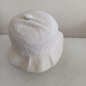 May include: A solid off-white bucket hat crafted from a textured, towel-like terry cloth fabric. The hat features a rounded crown with a small fabric-covered button at the very top center and a downward-sloping brim that encircles the entire head.