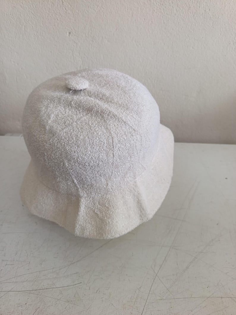 May include: A solid off-white bucket hat crafted from a textured, towel-like terry cloth fabric. The hat features a rounded crown with a small fabric-covered button at the very top center and a downward-sloping brim that encircles the entire head.