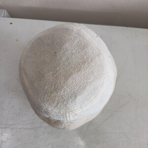 Pode incluir: Um gorro redondo de tecido texturizado em tom esbranqui&ccedil;ado, com topo plano e uma faixa lateral estruturada. O material tem uma apar&ecirc;ncia macia e ligeiramente felpuda, com costuras vis&iacute;veis que definem a coroa circular e o painel lateral.